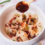 Gold spoon drizzling on chili oil onto wontons in a white bowl.