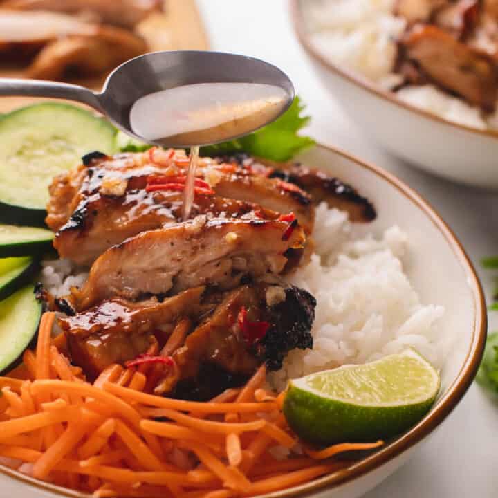 Silver spoon drizzling on nuoc cham sauce over lemongrass chicken rice bowl with cucumbers & carrots.