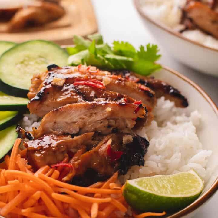 Rice bowl of lemongrass chicken with sliced cucumbers, carrots, and lime wedge.