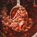 Silver spoon scooping chili oil with bits of garlic and chili from pan.