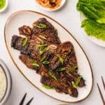 White oval plate with Kalbi topped with sesame seeds & green onions on a white table.