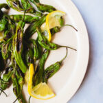 Charred shishito peppers with thin lemon slices on a white plate.