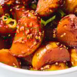 Sticky honey potatoes topped with sesame seeds and green onions in a white bowl.