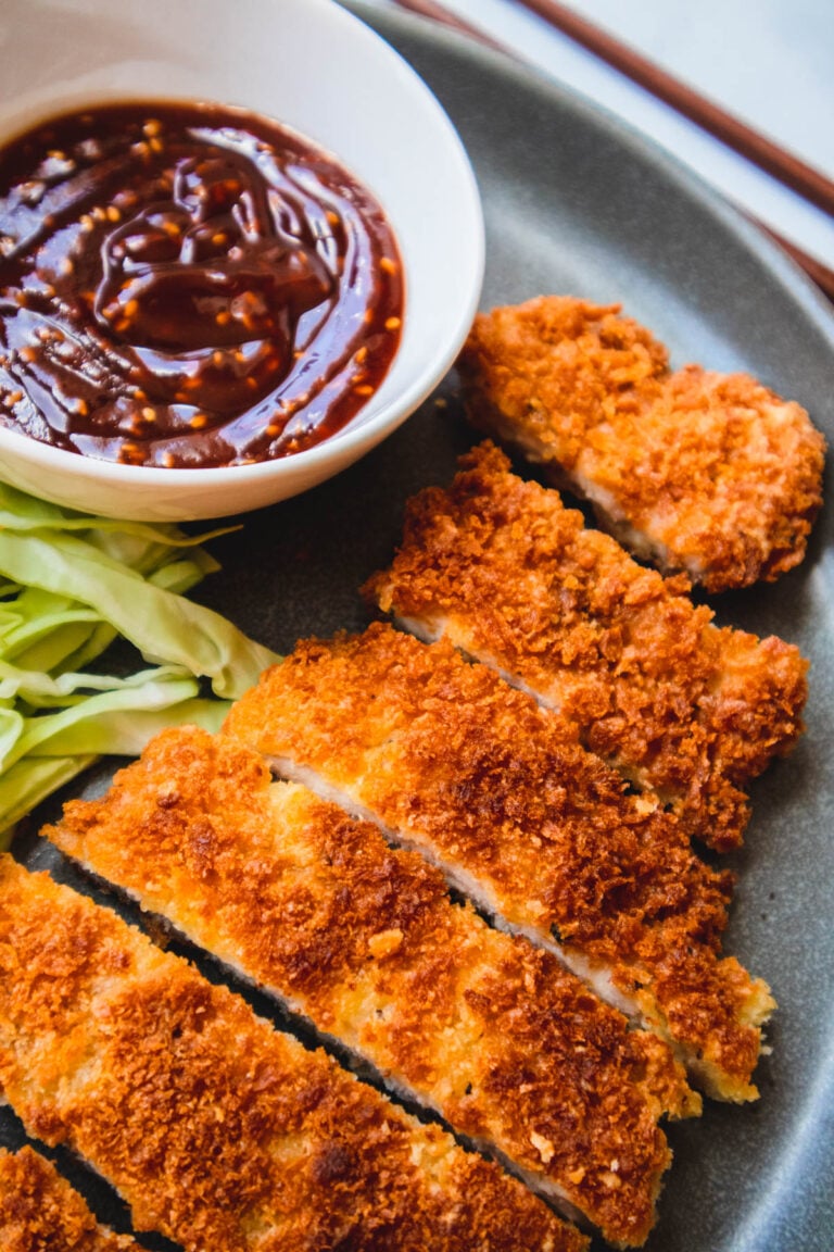 Crispy tonkatsu on a blue plate with shredded cabbage, dipping sauce, & chopsticks.