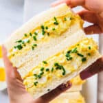 Holding two halves of Japanese egg salad sandwiches with chives sprinkled on top.