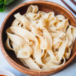 Boiled handmade noodles in a wood bowl next to bok choy.