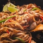 Chicken pad thai in a wok with a wooden spoon and lime wedges.