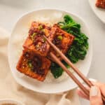 Picking up marinated tofu with chopsticks on a bed of rice and spinach.