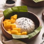 Mango sticky rice with black sesame seeds and mango chunks in a wooden bowl with a banana leaf.