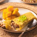 Adobo fried rice omelette on a white plate with silver spoon & fork on woven placemat.