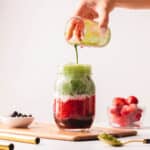 Hand pouring matcha into a glass mason jar of strawberry matcha latte on a wood plate.