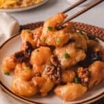Wooden chopstick picking up honey walnut shrimp from a white plate.