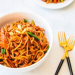 White serving bowl and plate of mie gorengs with gold forks on a white table.