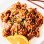 White plate of orange chicken topped with sesame seeds, green onions, & orange slices.