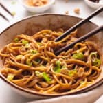 Peanut noodles in a white bowl with chopsticks and topped with green onions and crushed peanuts.