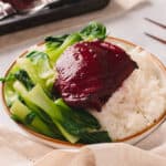 White bowl of rice with char siu chicken slices and bok choy on a white table.