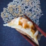 Wooden chopsticks holding gyoza with a crispy skirt against a blue background.