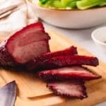 Sliced char siu pork on a wooden cutting board with a bowl of bok choy in the back.