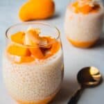 Glass cups of mango sago topped with mango and coconut on a white table.