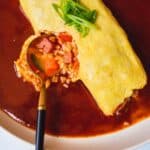 Plate of omurice with sauce and gold spoon cutting inside omelette.