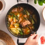 A silver spoon scooping pork from a pot of sinigang na baboy on a white table with vegetables.