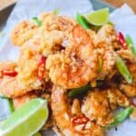 A plate of crispy szechuan shrimp topped with chilies, green onions, & lime wedges on side.