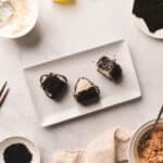 White rectangle plate with three tuna onigiri with black sesame seeds on a white table.