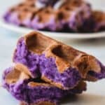 Torn ube mochi waffles laying on a white table with a plate of waffles in back.