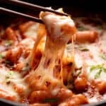Wooden chopsticks pulling cheesy tteokbokki from a black pan.