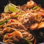 Shrimp pad thai with lime wedges and crushed peanuts in a wok.