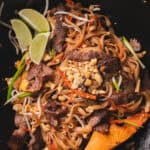 Beef pad thai topped with crushed peanuts and lime wedges in a wok.