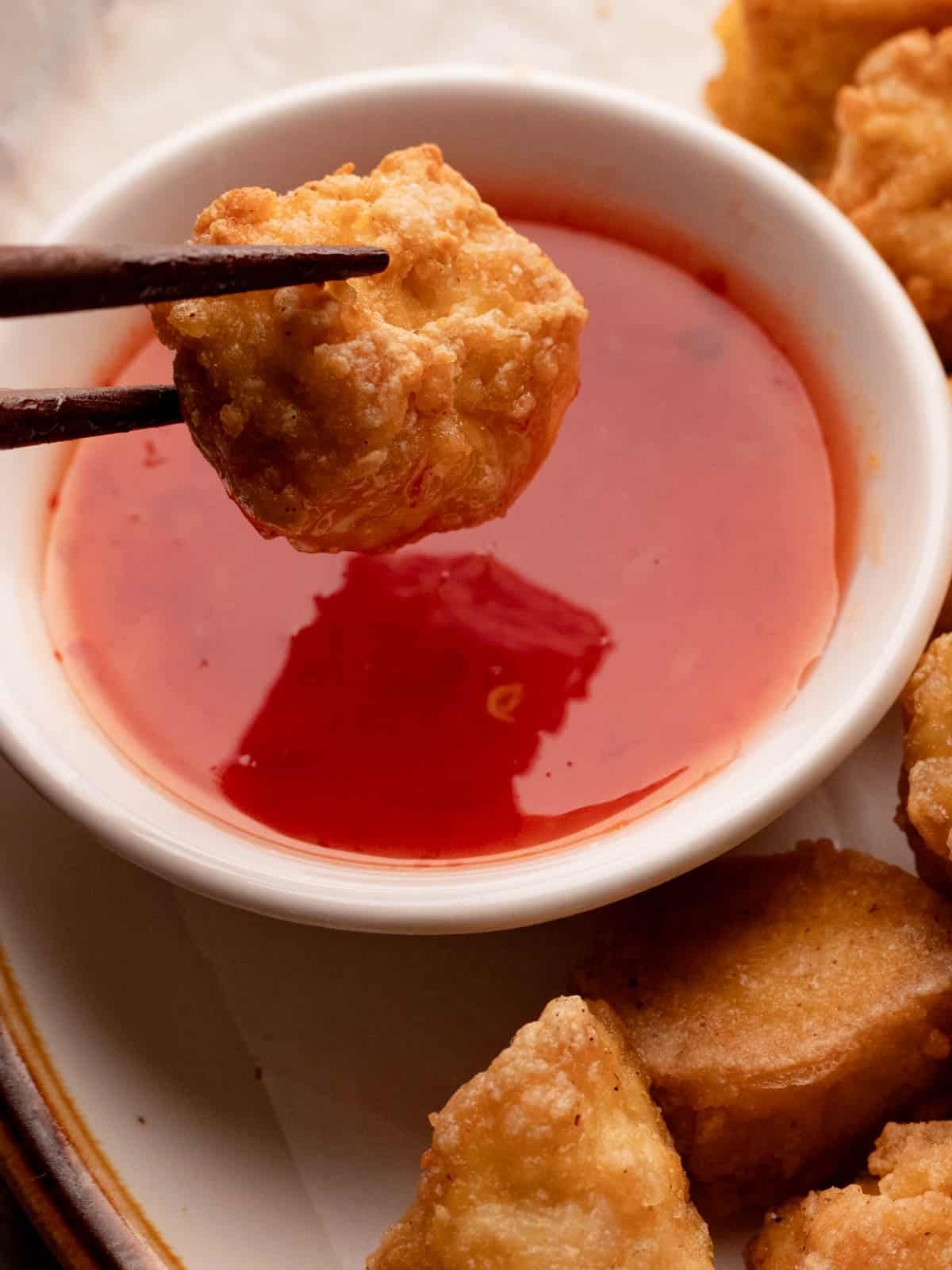 Crispy golden tofu held with wooden chopsticks over sweet chili sauce.