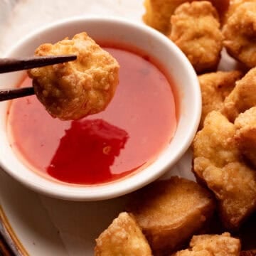 Crispy golden tofu held with wooden chopsticks over sweet chili sauce.