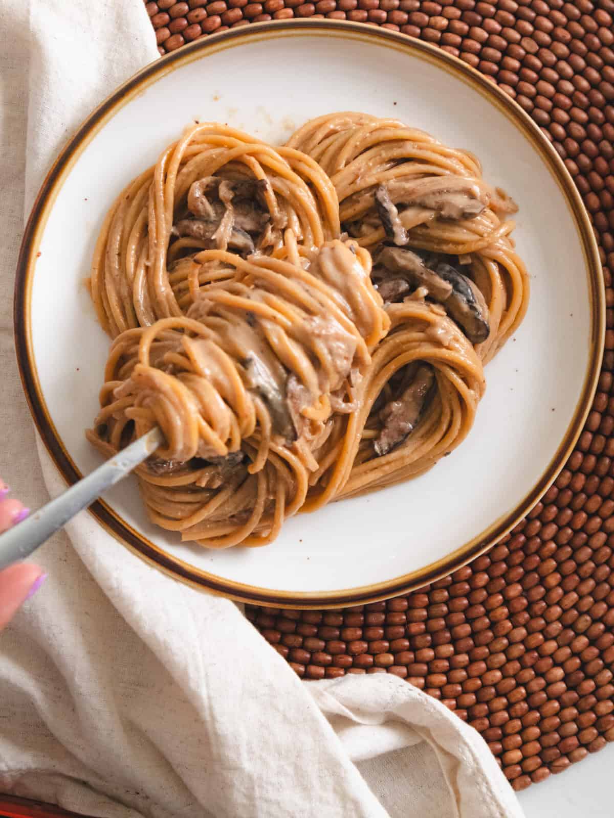 Fork twirling mushroom miso pasta on a white brown rimmed plate on a beaded placemat.