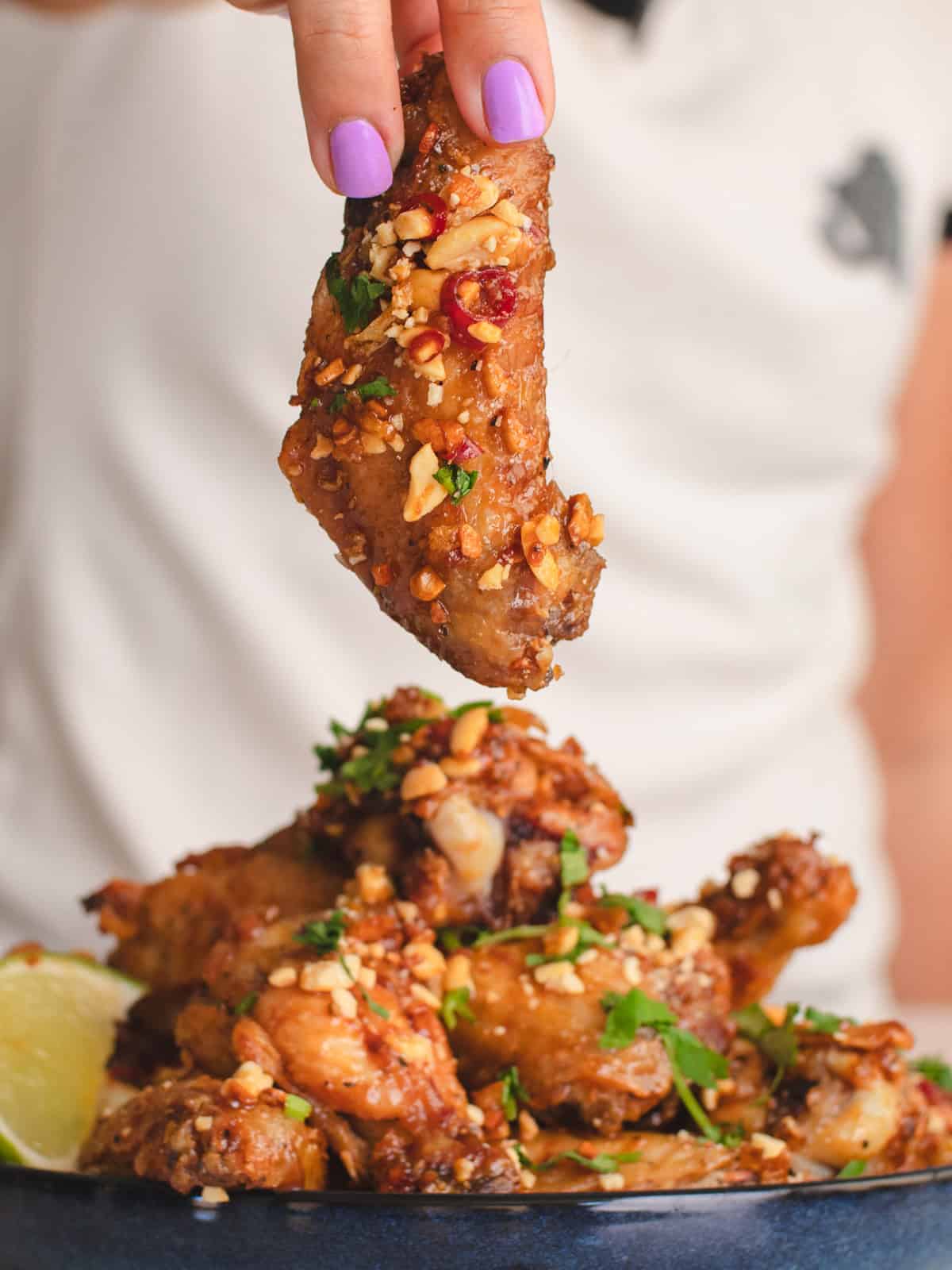Holding a tamarind chicken wing coated in crushed peanuts, fried shallots, & crispy garlic over a plate of wings.