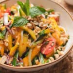 Green mango salad in a brown rimmed white bowl garnished with herbs & crushed peanuts.