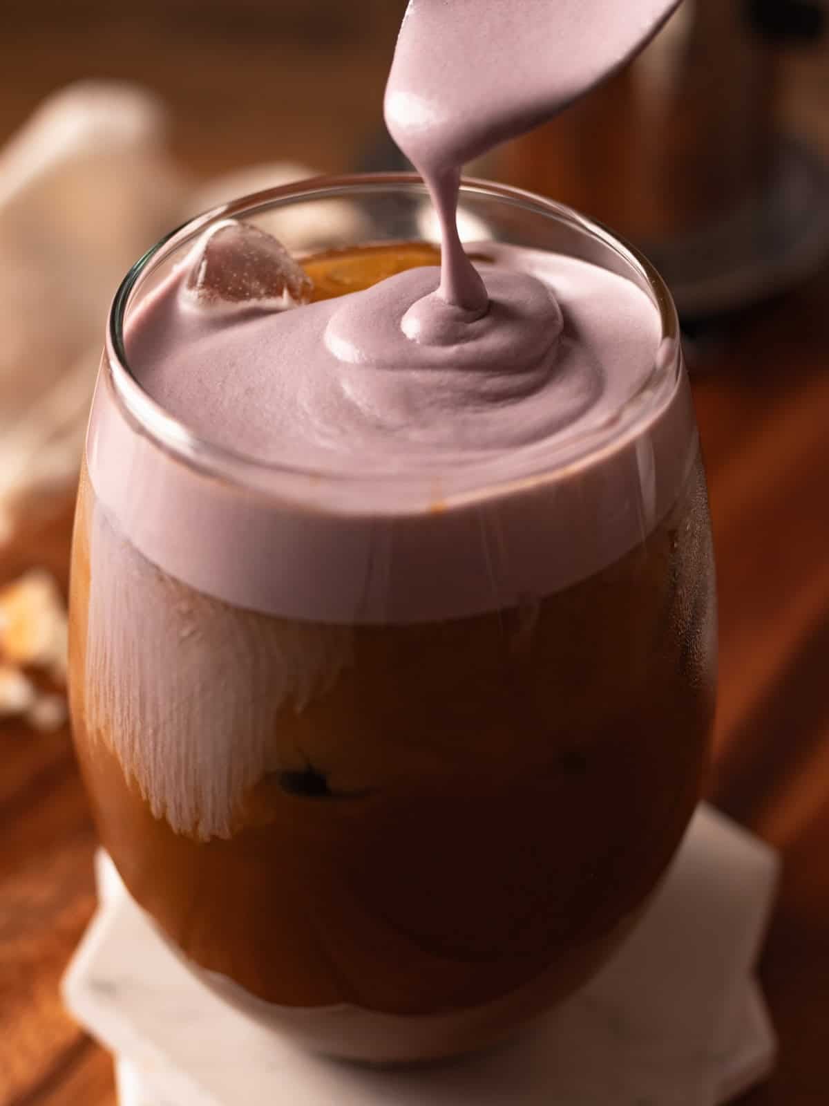 Silver spoon dripping ube cream foam on a glass of iced coffee.
