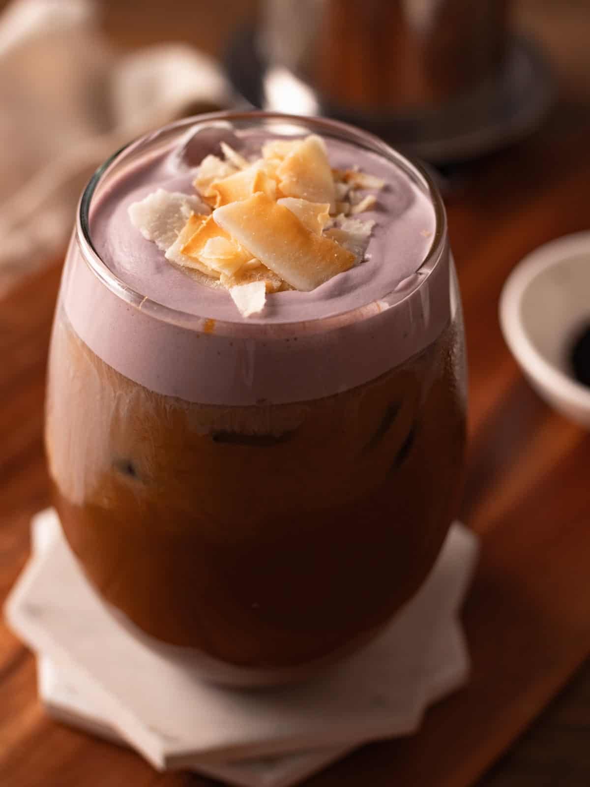 Glass cup of ube latte garnished with toasted coconut chips on white coasters.