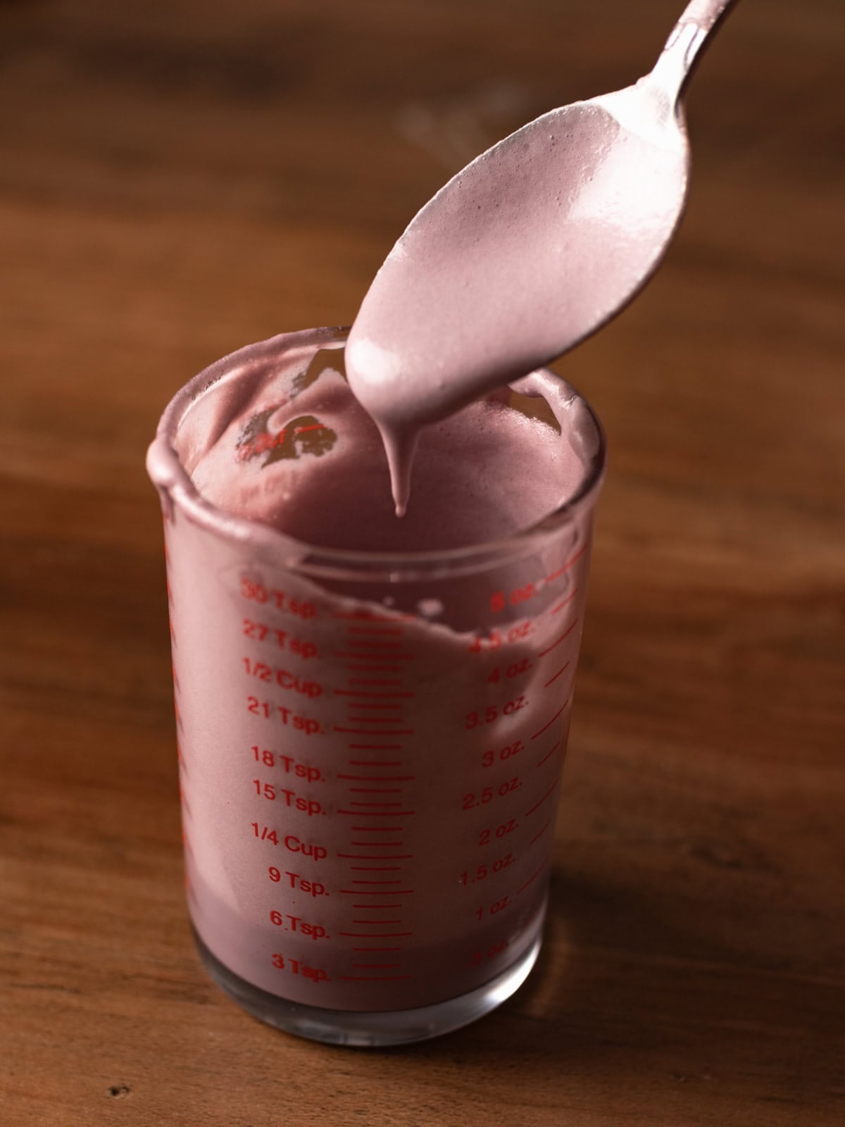 Silver spoon dripping ube foam dripping in a glass measuring cup.