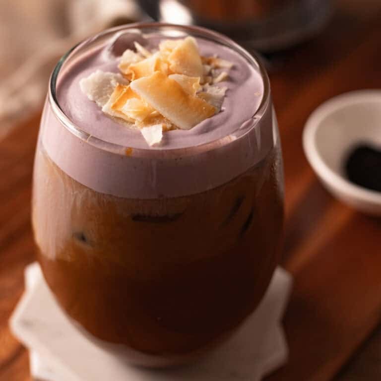 Glass cup of ube latte garnished with toasted coconut chips on white coasters.