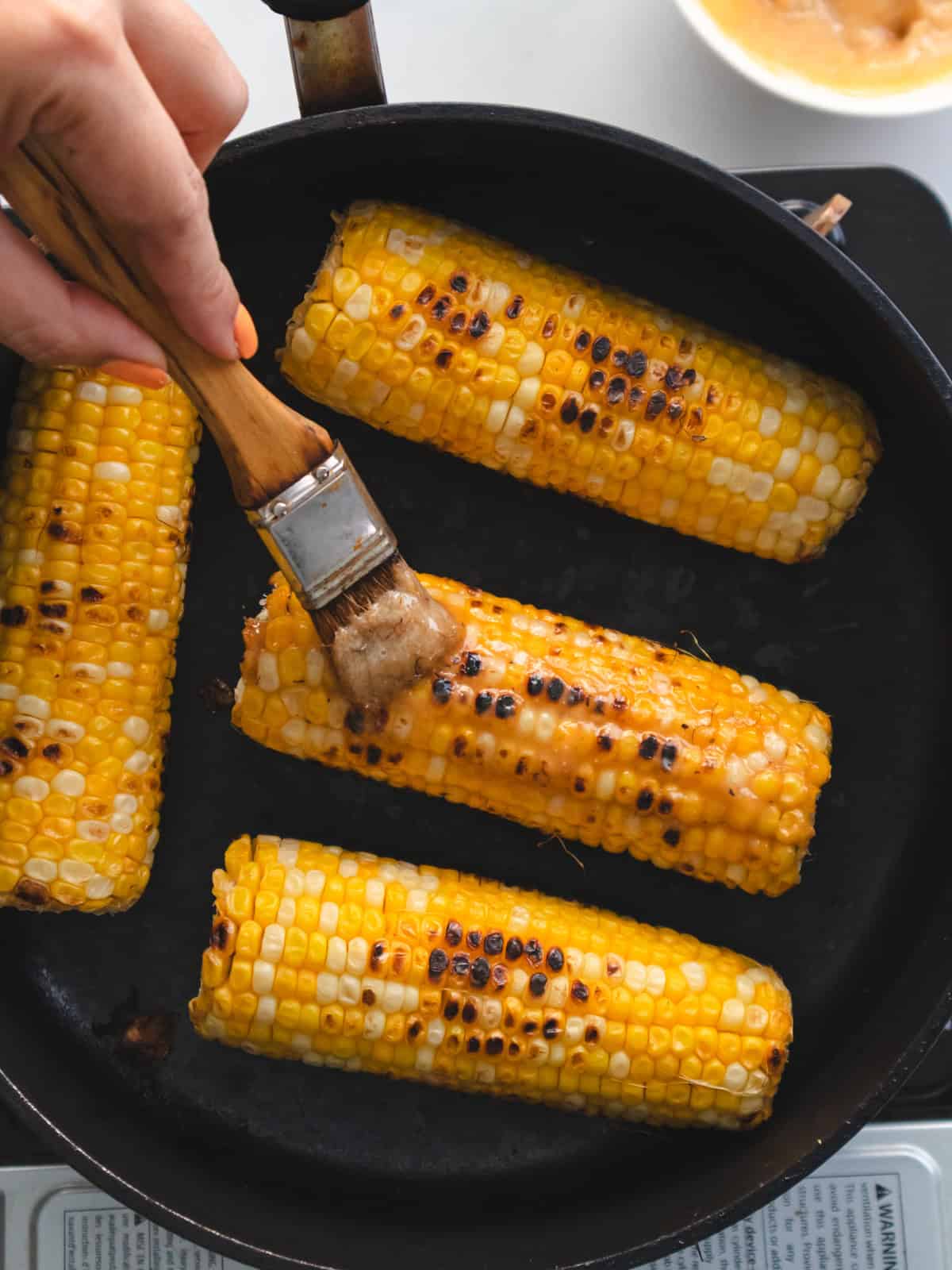 Charred corn cobs cooking stovetop on a skillet being brushed with miso butter.